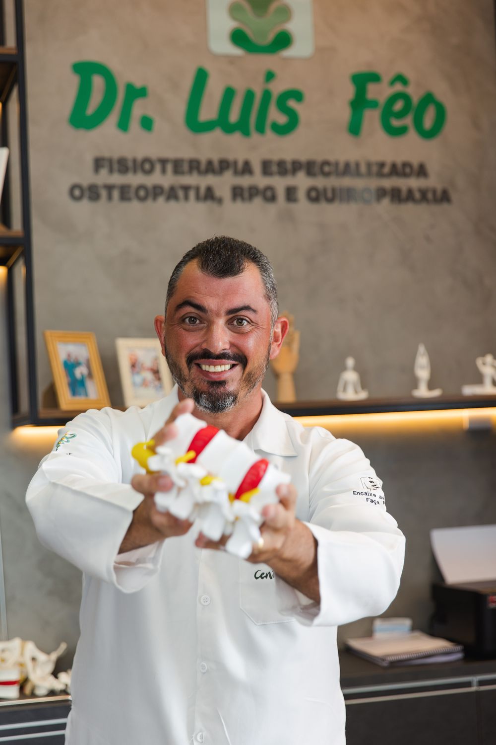 Dr. Luis Fêo no consultório, segurando um modelo anatômico de vértebras lombares